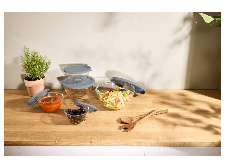 Bols en verre avec couvercles gris, remplis de salade, soupe et olives, sur un comptoir en bois.