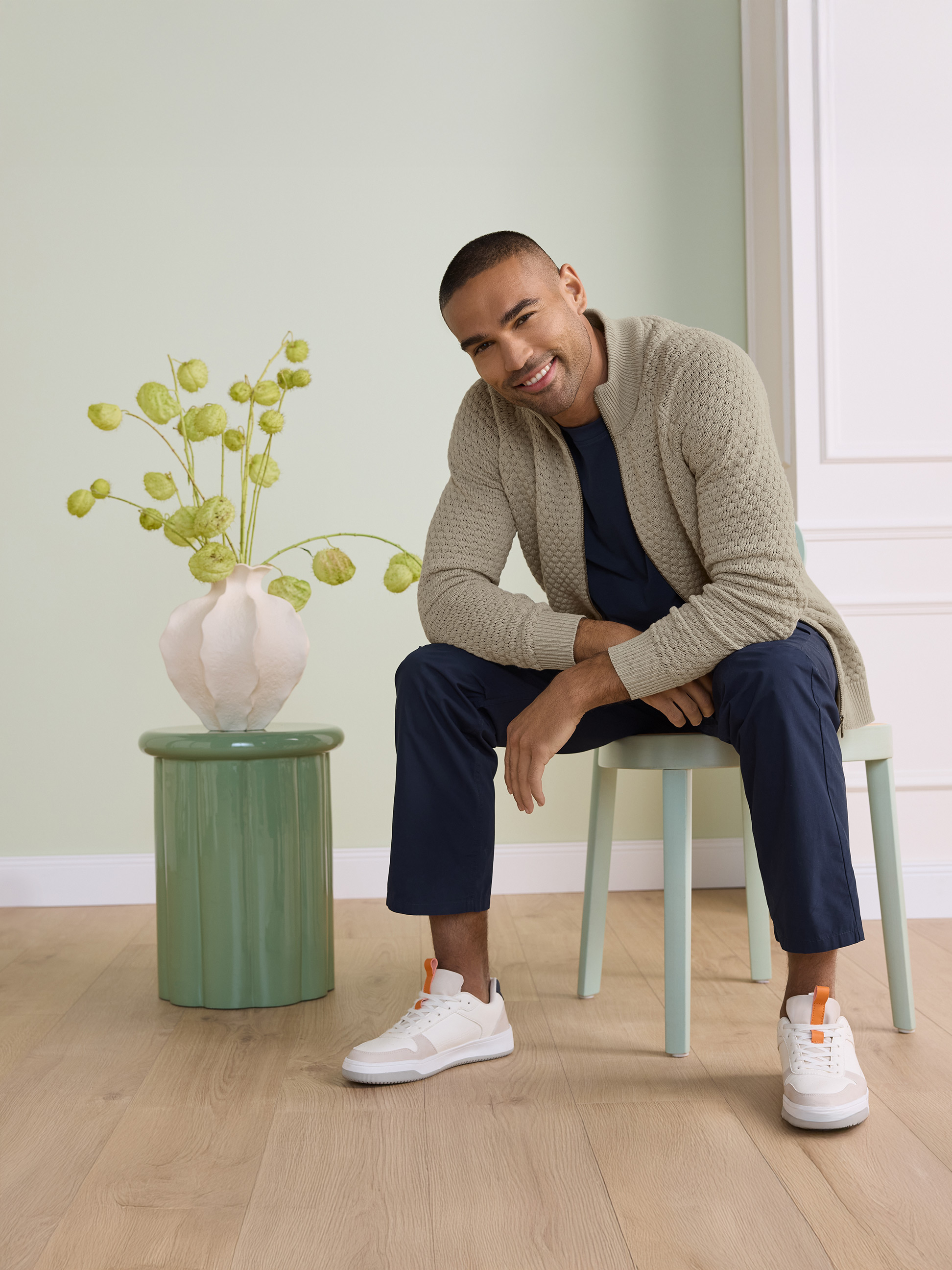 Homme souriant en cardigan beige, t-shirt bleu marine et pantalon, assis sur une chaise.