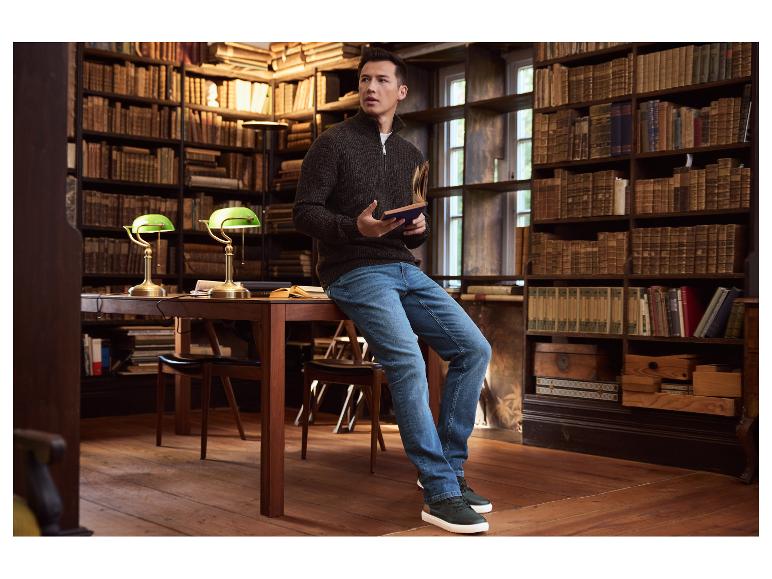 Homme en pull marron et jean assis sur une table dans une bibliothèque, tenant un livre.