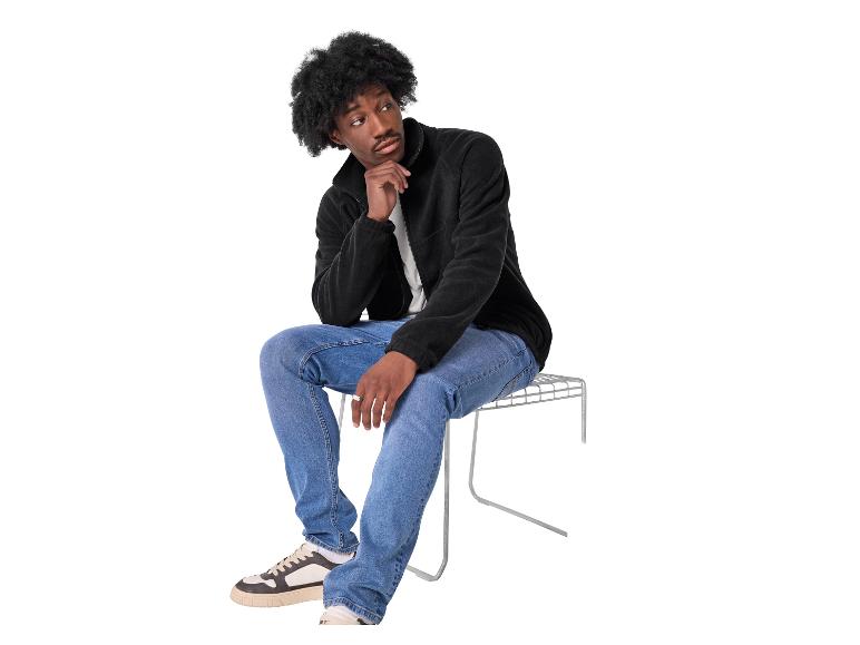 Un homme en veste polaire noire et jean bleu est assis sur un tabouret en métal, l'air pensif.