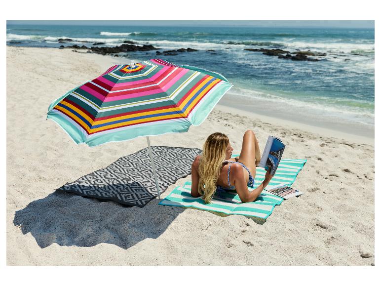 Femme lisant sur un tapis de plage CRIVIT sous un parasol coloré sur la plage.