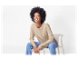 Femme souriante portant un pull beige et un jean, assise sur une chaise.