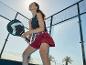 Vrouw in zwart sporttopje en rode rok met padelracket op een padelbaan.