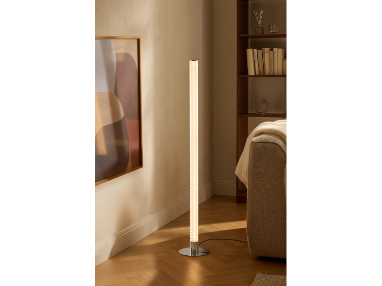 Cilindervormige vloerlamp met chromen voet verlicht een moderne woonkamer met boekenplank en bank.