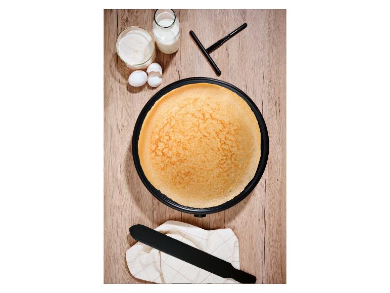 Bovenaanzicht van een crêpe-ijzer met een gouden crêpe, omringd door bloem, melk en eieren.