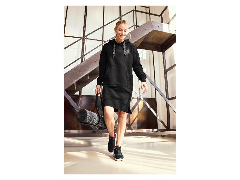 Een vrouw in een zwarte hoodie-jurk en zwarte sneakers met een yogamat.