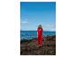 Femme en combinaison rouge sur une côte rocheuse avec la mer et le ciel bleu en arrière-plan.