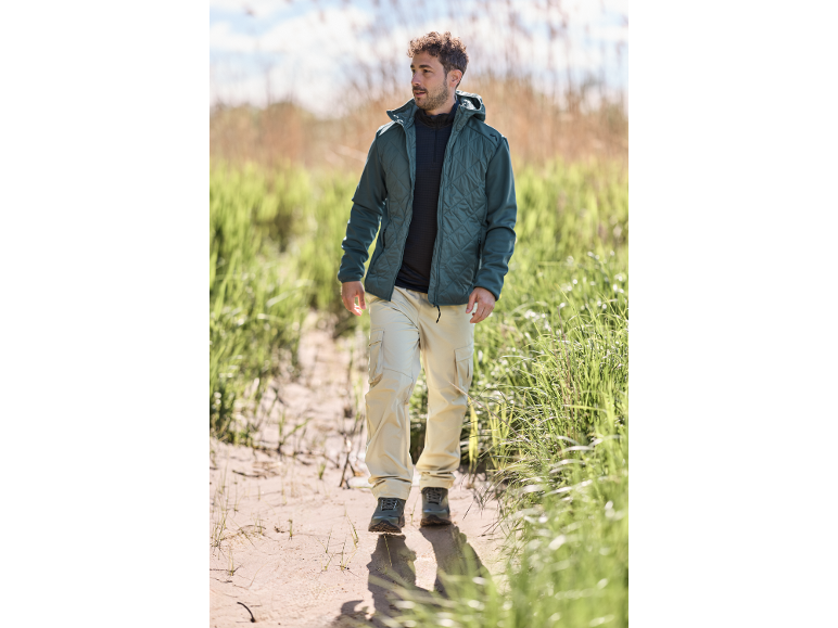 Man in gewatteerde jas en beige cargobroek loopt over een zandpad.