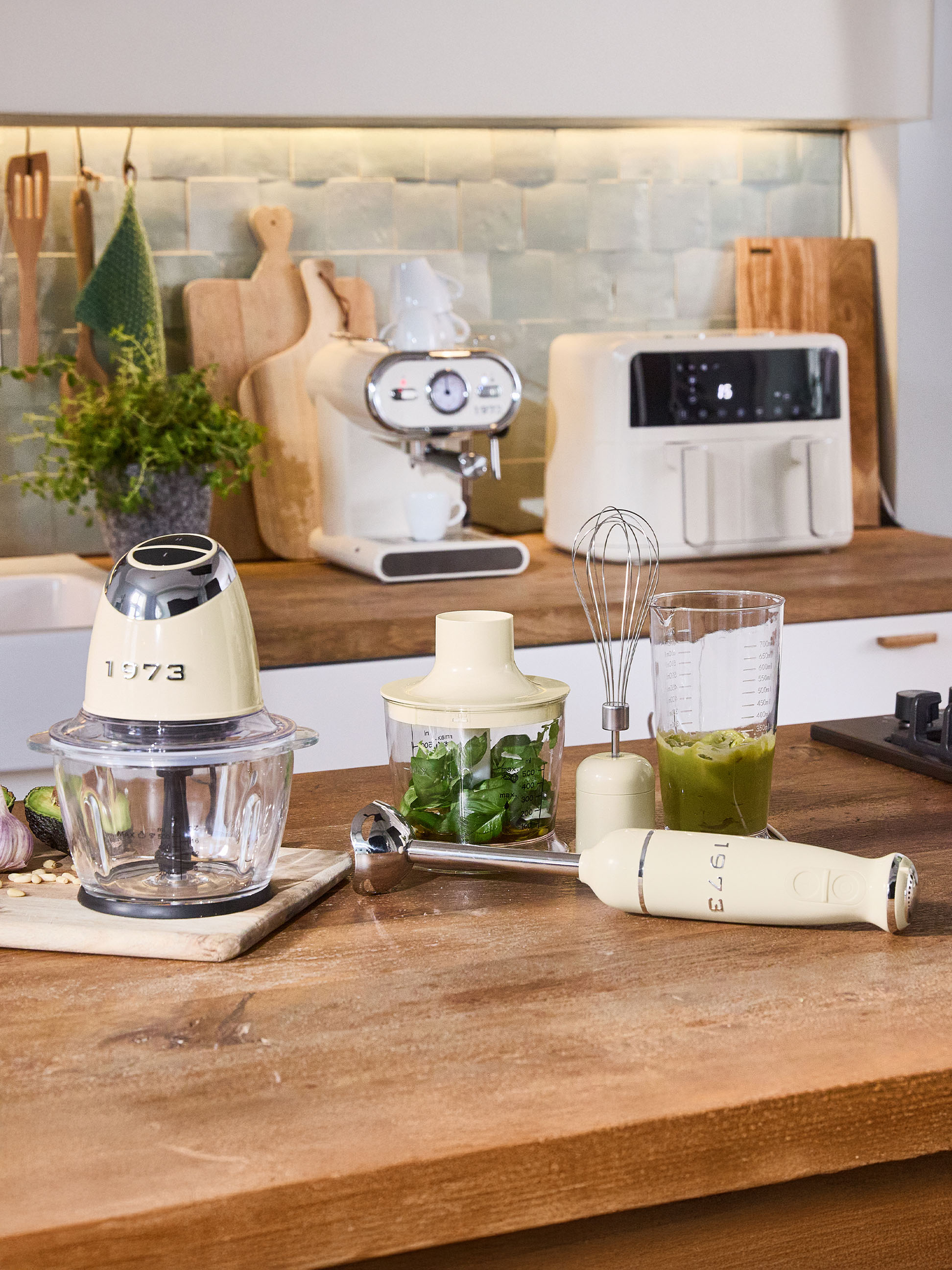 Crèmekleurige keukenapparatuur, waaronder een hakmolen, staafmixer en espressomachine, op een houten aanrecht.