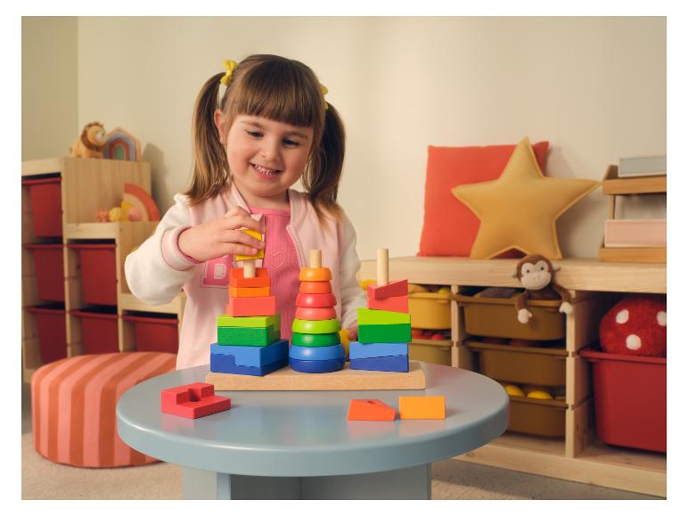 Lachend meisje speelt met kleurrijk houten stapelspeelgoed op een grijze tafel in een speelkamer.