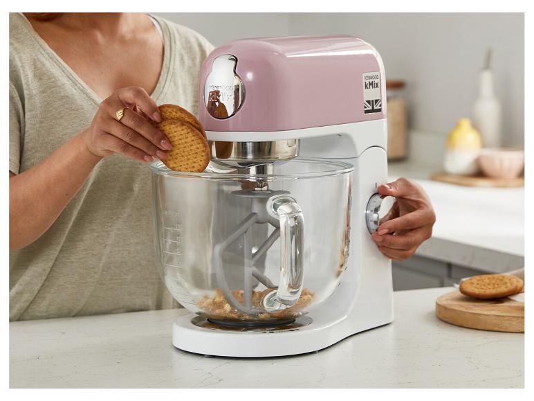 Une femme met des biscuits dans un robot pâtissier rose et blanc sur un comptoir de cuisine.