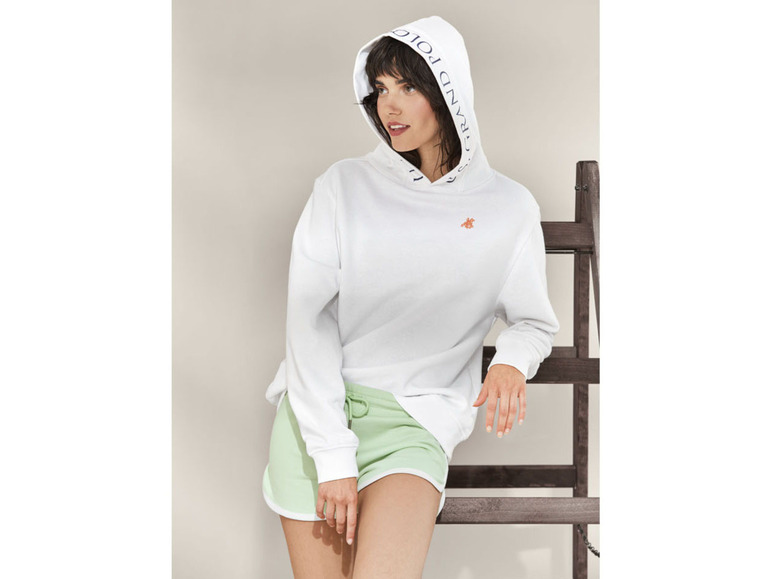 Een vrouw draagt een witte hoodie met de tekst 'Grand Polo' en groene shorts.