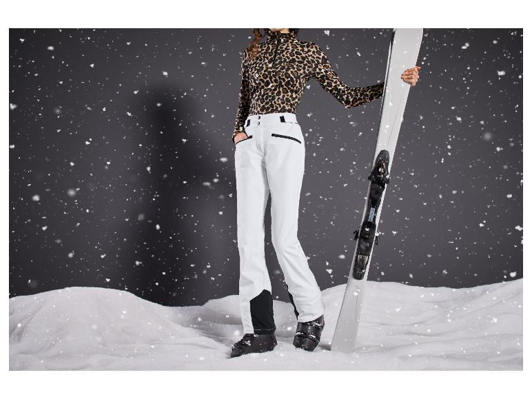 Une femme portant un pantalon de ski blanc, des chaussures de ski noires et un haut léopard tient des skis blancs.