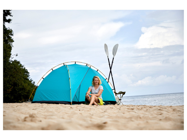 Une femme assise devant une tente bleue sur la plage avec deux pagaies à côté.