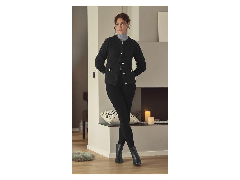 Vrouw in een zwart vest en zwarte legging, met laarzen, bij een open haard.