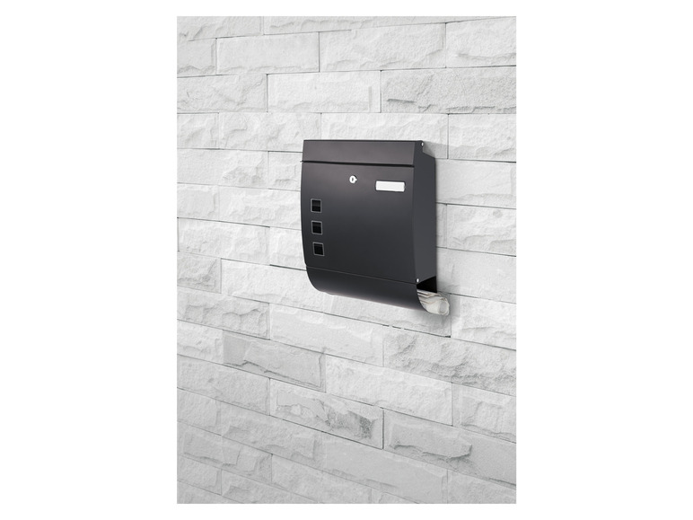 Een zwarte brievenbus met een krantenbak aan de zijkant, gemonteerd op een witte bakstenen muur.