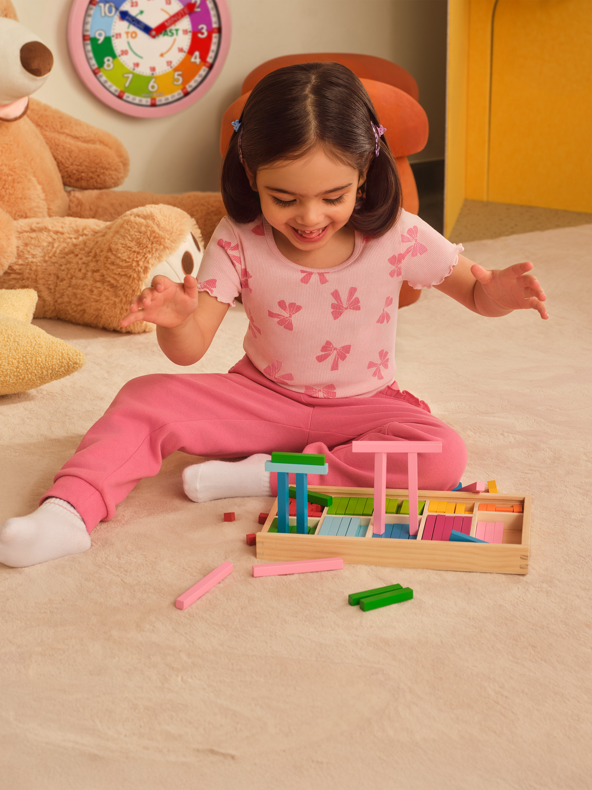 Lachend meisje speelt met kleurrijke houten blokken op een tapijt, met een teddybeer en klok op de achtergrond.