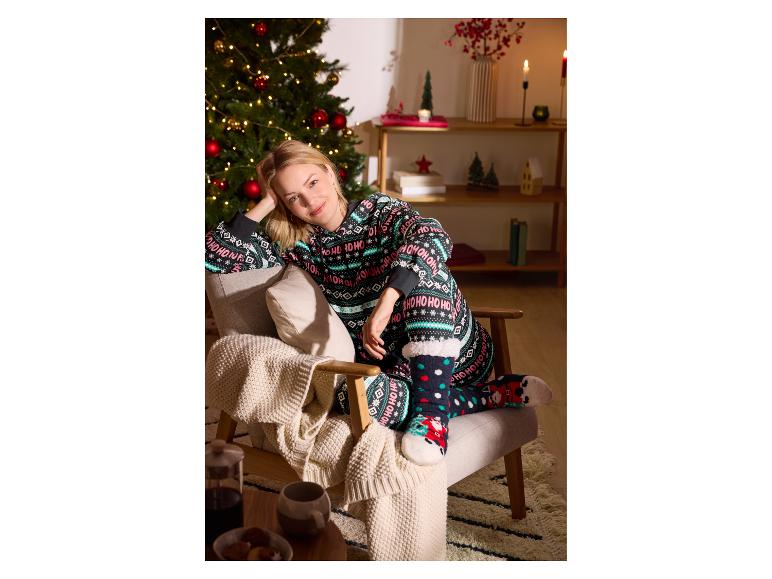 Vrouw in kerstpyjama met „Ho Ho Ho“-patroon en sokken, zittend in een fauteuil naast een kerstboom.