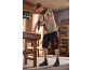 Man in zwarte werkshorts en beige T-shirt gebruikt een boormachine op een werkbank.