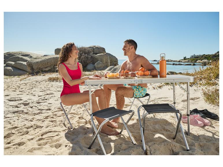 Couple pique-niquant sur la plage avec table et chaises pliantes, fruits et bouteille d'eau.
