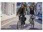 Een stel fietst door een stadstraat met gebouwen en een blauwe auto op de achtergrond.