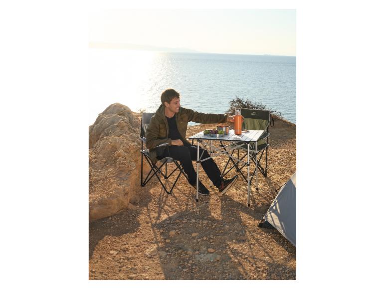 Homme assis à une table de camping avec des chaises, à côté d'une tente, face à la mer.