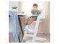 Baby in witte houten kinderstoel aan tafel met blauwe deken en plant.