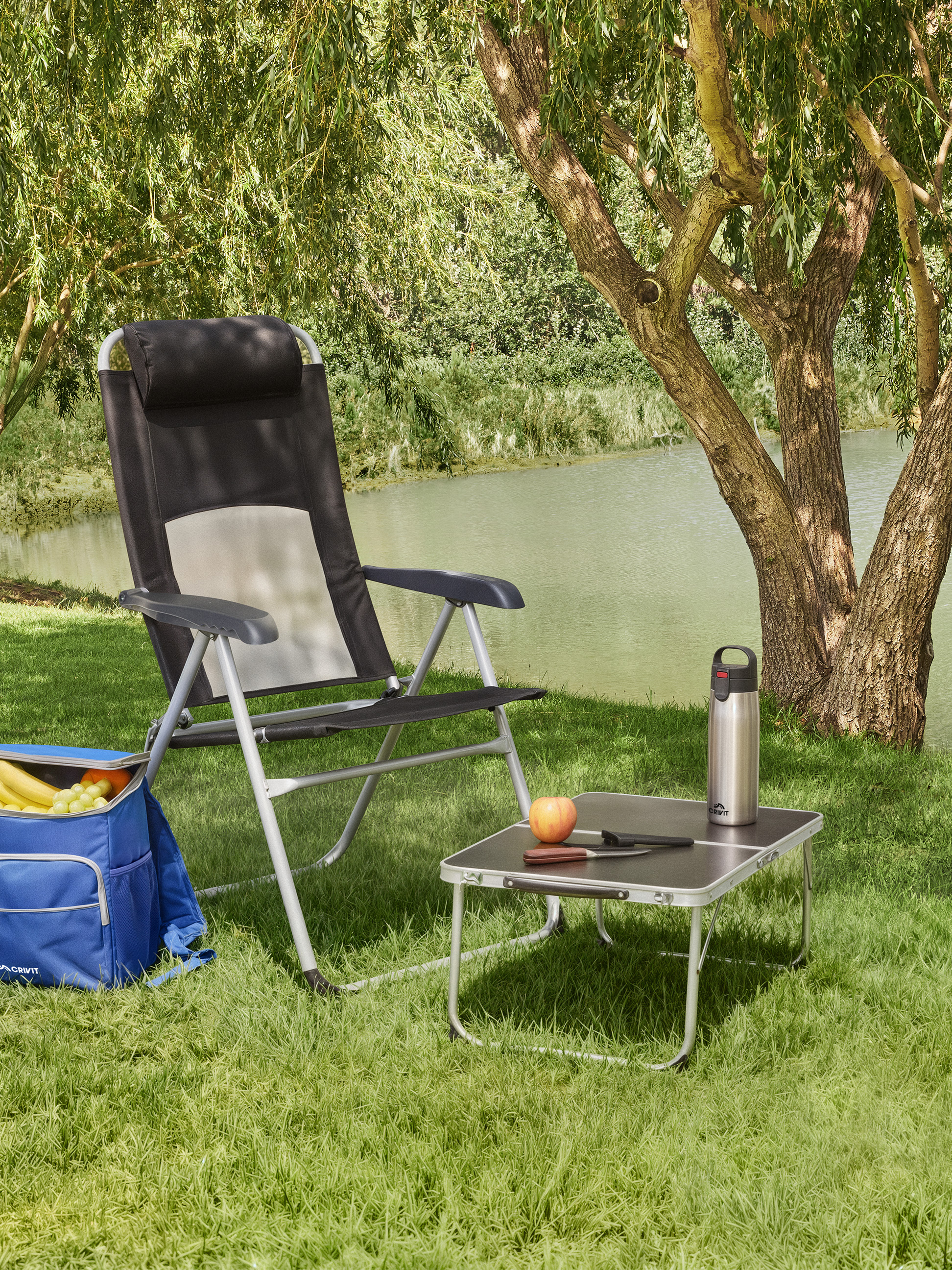 Opvouwbare campingstoel, kleine tafel, koeltas en waterfles bij een meer.