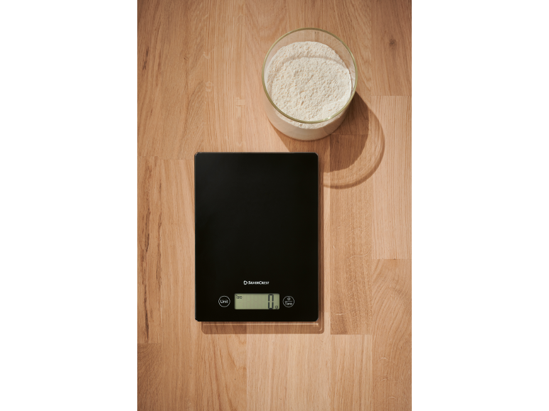 Une balance de cuisine noire affichant '0 g' à côté d'un bol en verre de farine sur une surface en bois.