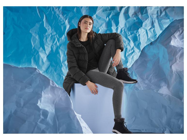 Femme en veste noire et leggings gris, assise sur un cube bleu, sur fond bleu.