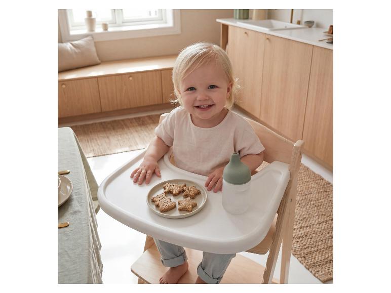 Jeune enfant souriant dans une chaise haute en bois avec des biscuits et un biberon.