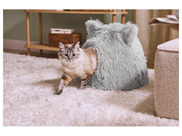 Een kat komt uit een pluizig, zacht bed op een wit tapijt, naast een houten rek.