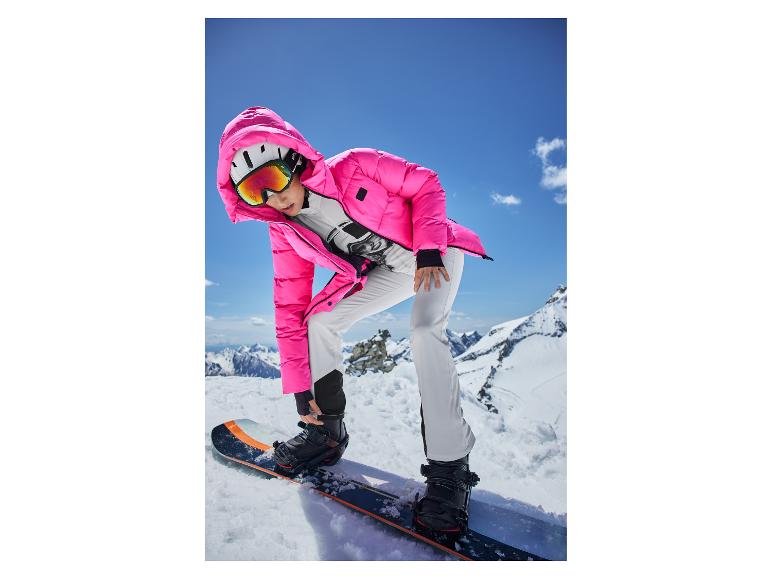 Une femme en veste rose et pantalon de snowboard blanc, ajustant les fixations sur un snowboard.