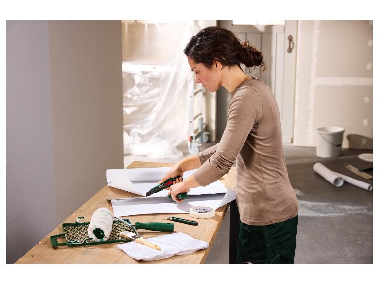 Vrouw snijdt behang met een stanleymes, met schildersbenodigdheden op tafel.