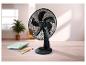 Ventilateur de bureau noir sur un bureau en bois avec cahiers et stylos, dans une pièce lumineuse.