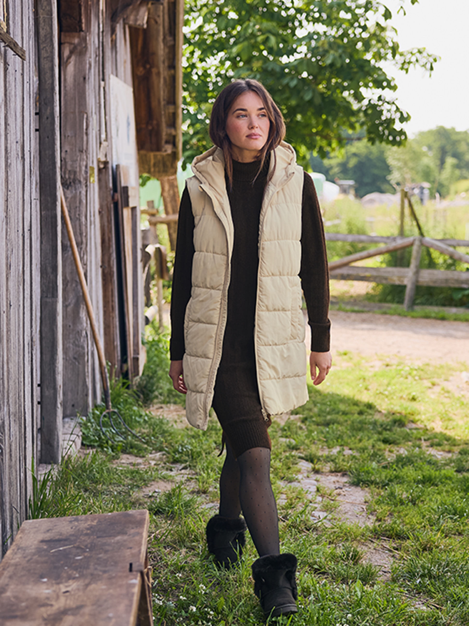 Vrouw in een beige bodywarmer en bruine jurk, lopend op een landweg.