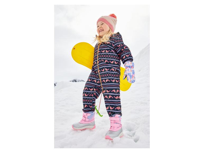 Fille souriante en combinaison de neige à motifs et bottes roses, tenant une luge jaune.