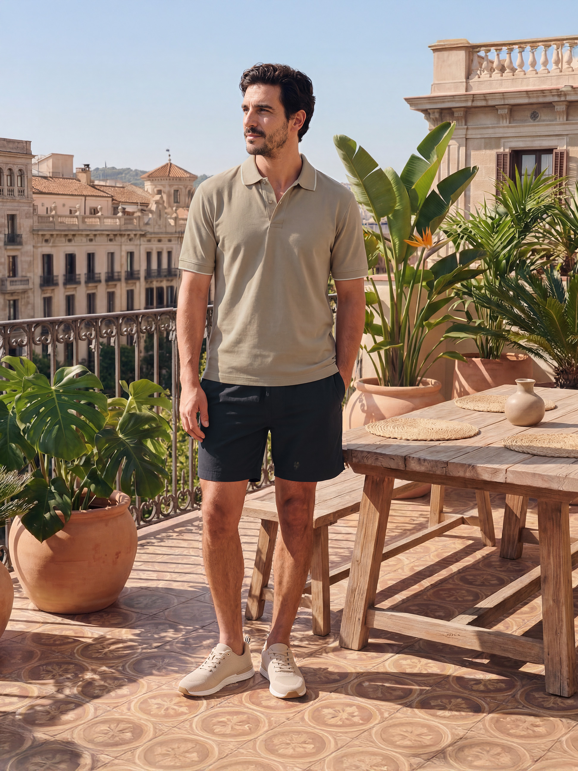 Man in beige poloshirt en zwarte korte broek op zonnig terras met uitzicht op de stad.