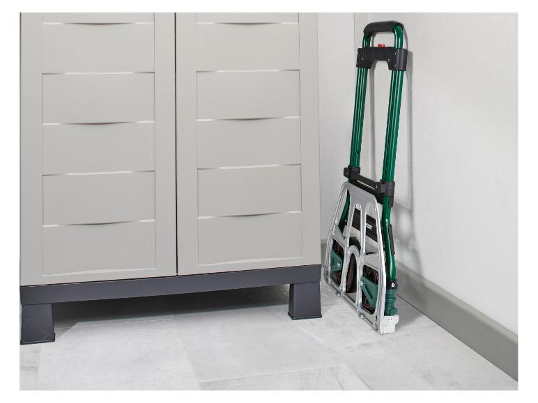 Een opgevouwen groene en zilveren steekwagen leunt tegen een witte muur naast een lichtgrijze opbergkast.