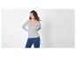 Vrouw in een lichtgrijze top met lange mouwen en blauwe jeans, opzij kijkend.