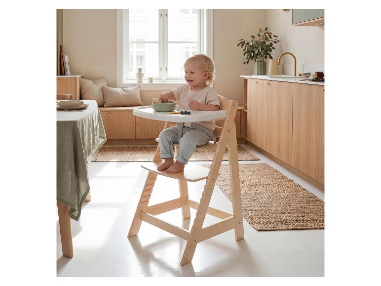Bébé souriant dans une chaise haute en bois, mangeant des biscuits et des myrtilles dans une cuisine lumineuse.
