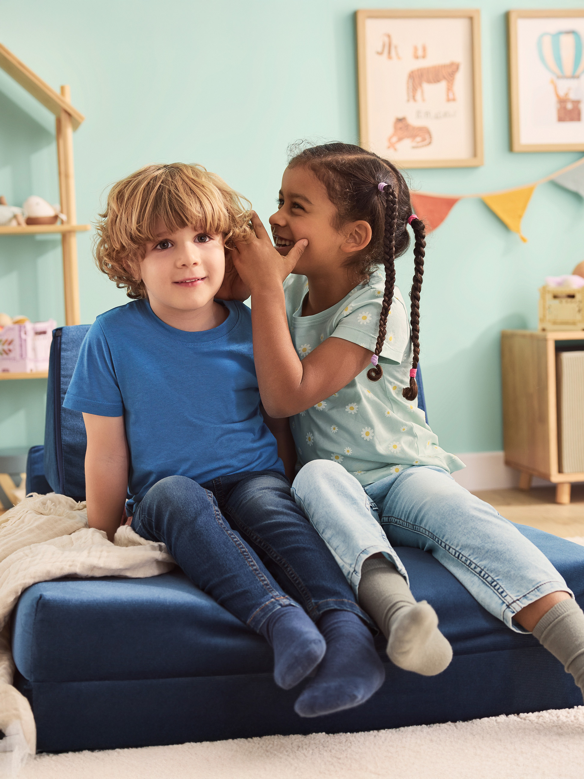 Twee kinderen zitten op een blauwe opvouwbare bank, de een fluistert de ander in het oor.