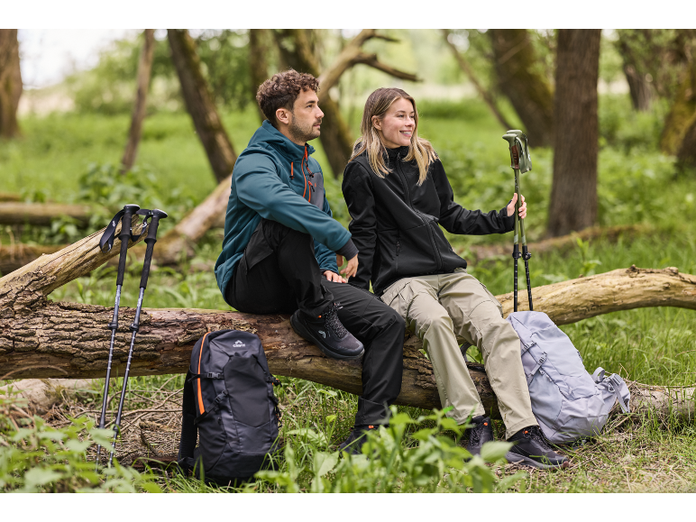 Koppel in wandelkleding zit op boomstam met rugzakken en trekkingstokken.