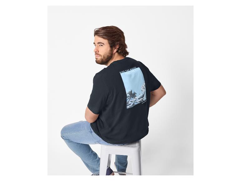 Man in zwart T-shirt met strandprint en jeans, zittend op een kruk.