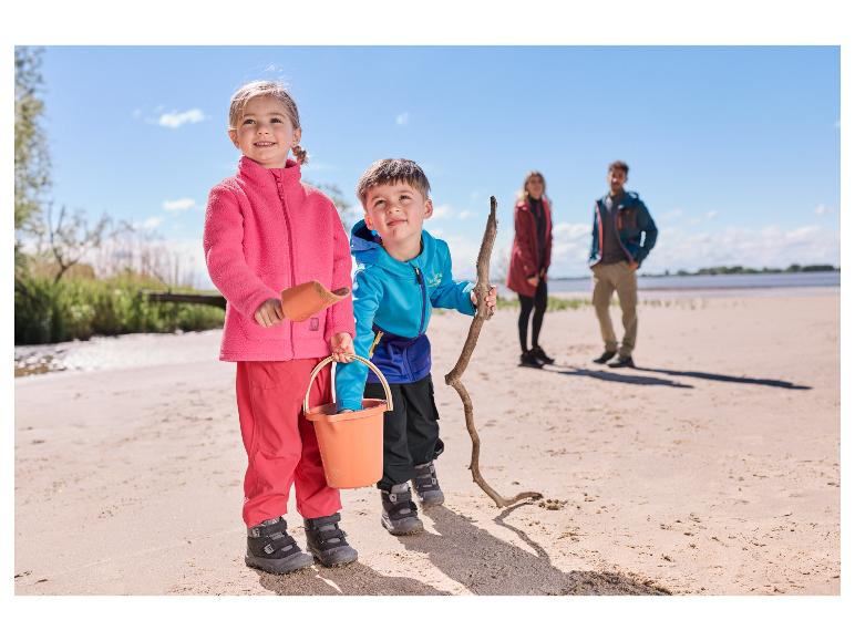 Deux enfants en vestes softshell LUPILU® jouant sur une plage de sable avec leurs parents en arrière-plan.