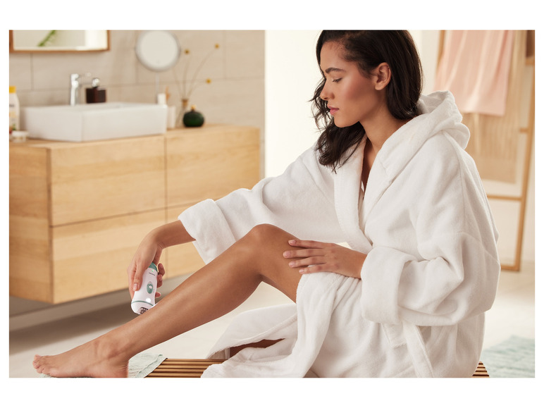 Une femme en robe de chambre utilise un épilateur Braun sur ses jambes.