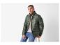 Man in een groene pufferjas en blauwe jeans, zittend op een kruk.