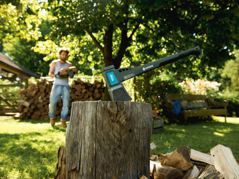 Une hache Gardena dans une souche d'arbre avec du bois de chauffage en arrière-plan.