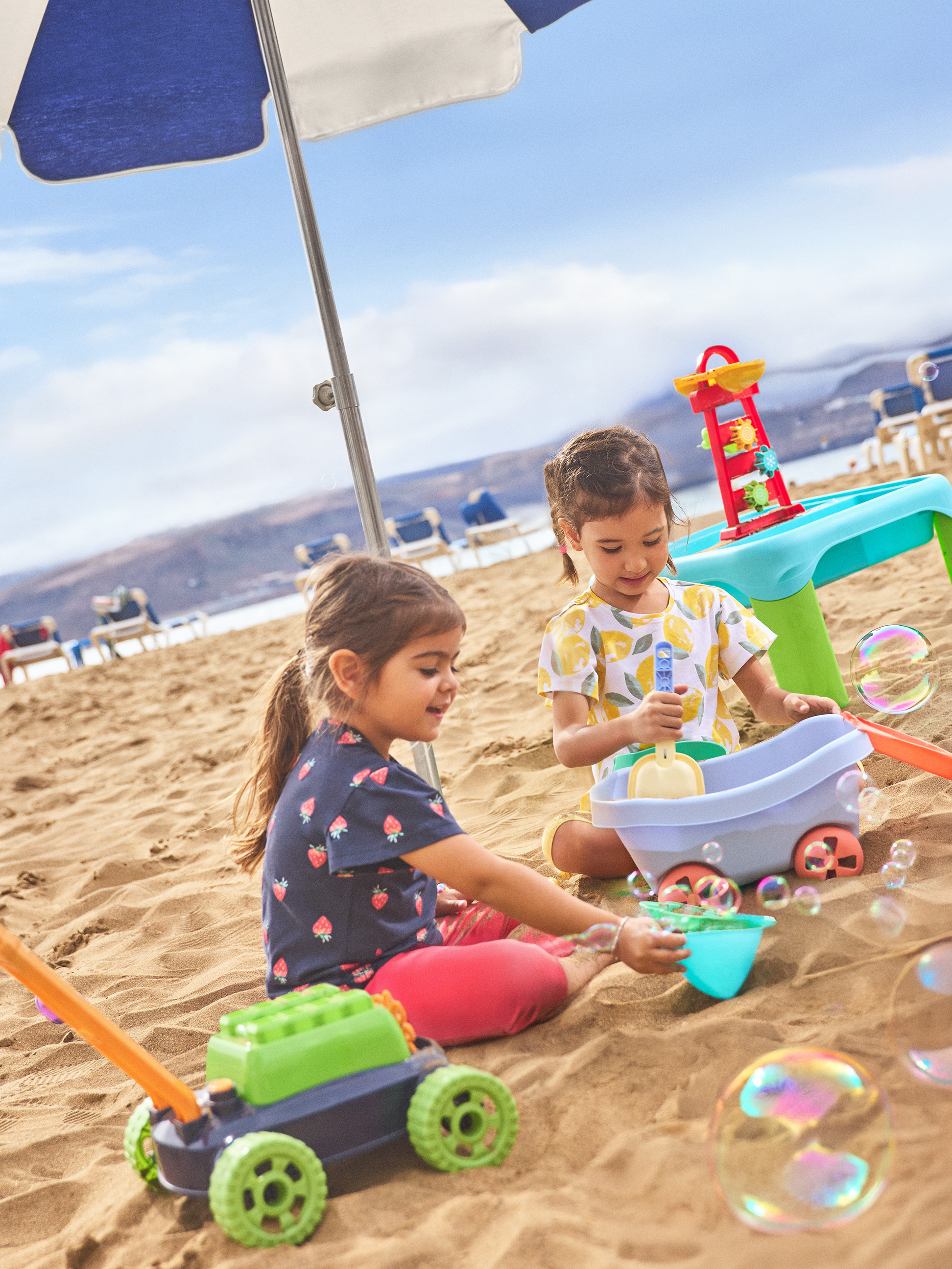 Twee kleine meisjes spelen met strandspeelgoed en zeepbellen onder een parasol.