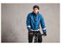 Man in blauwe fietsjas met reflecterende strepen, fietshelm en zwarte handschoenen op een zwarte fiets.